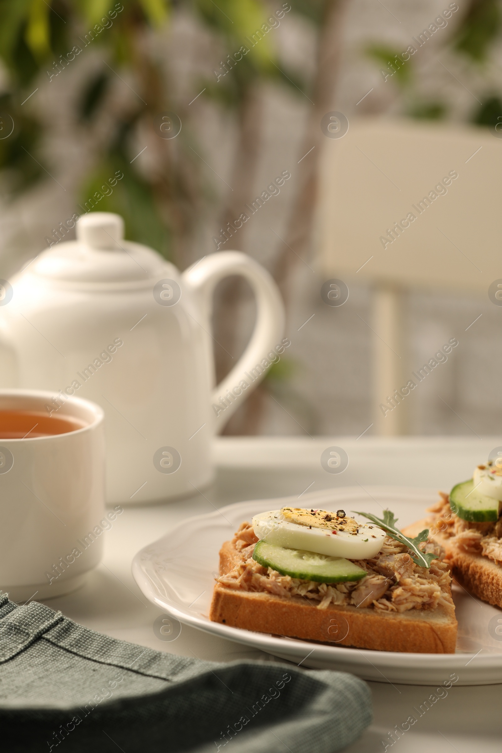 Photo of Delicious sandwiches with tuna, vegetables, boiled egg and cup of tea on white table indoors, closeup. Tasty breakfast