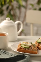 Delicious sandwiches with tuna, vegetables, boiled egg and cup of tea on white table indoors, closeup. Tasty breakfast