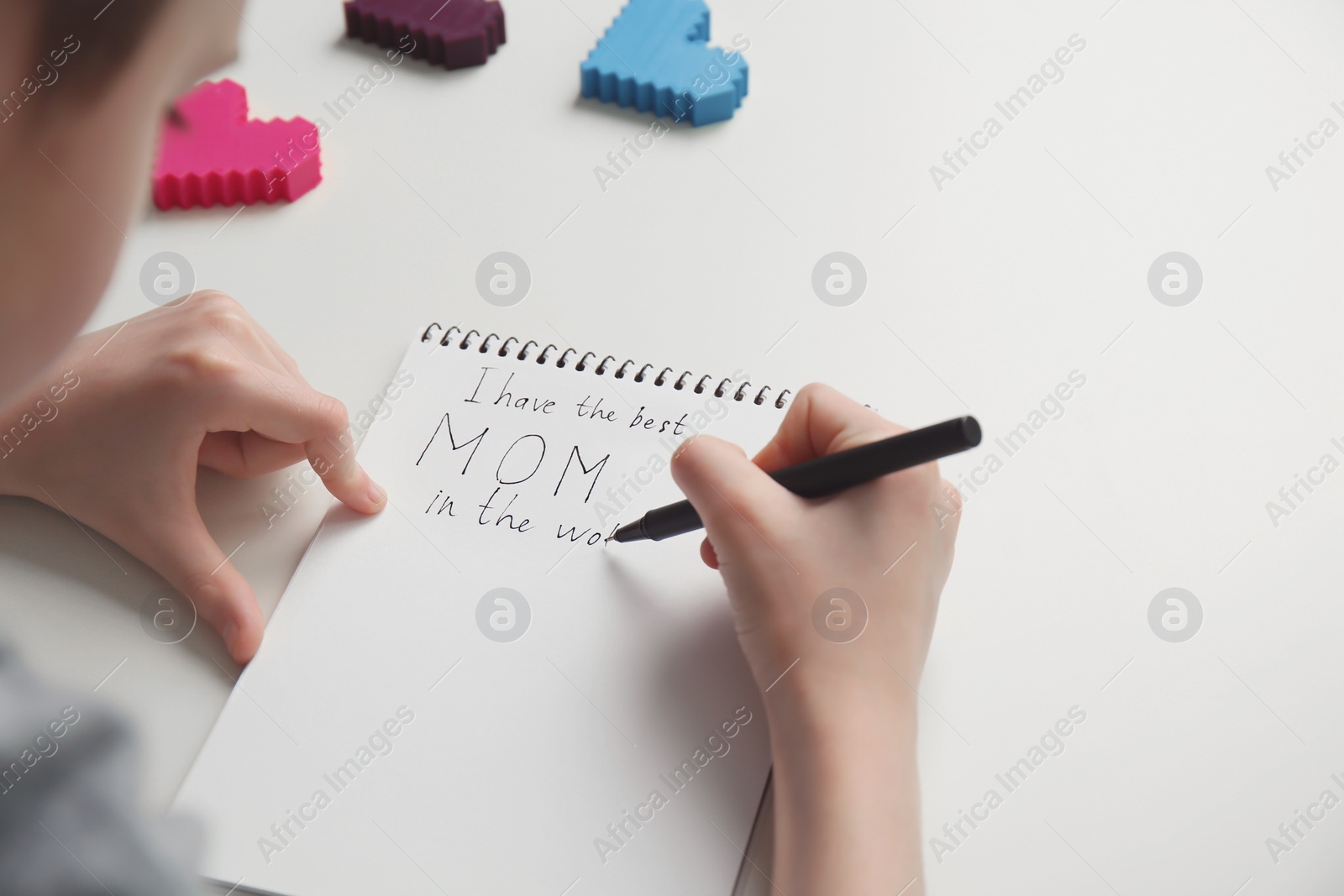 Photo of Little child writing congratulation for Mother's Day in notebook on light background