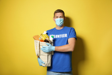 Photo of Male volunteer in protective mask and gloves with products on yellow background. Aid during coronavirus quarantine