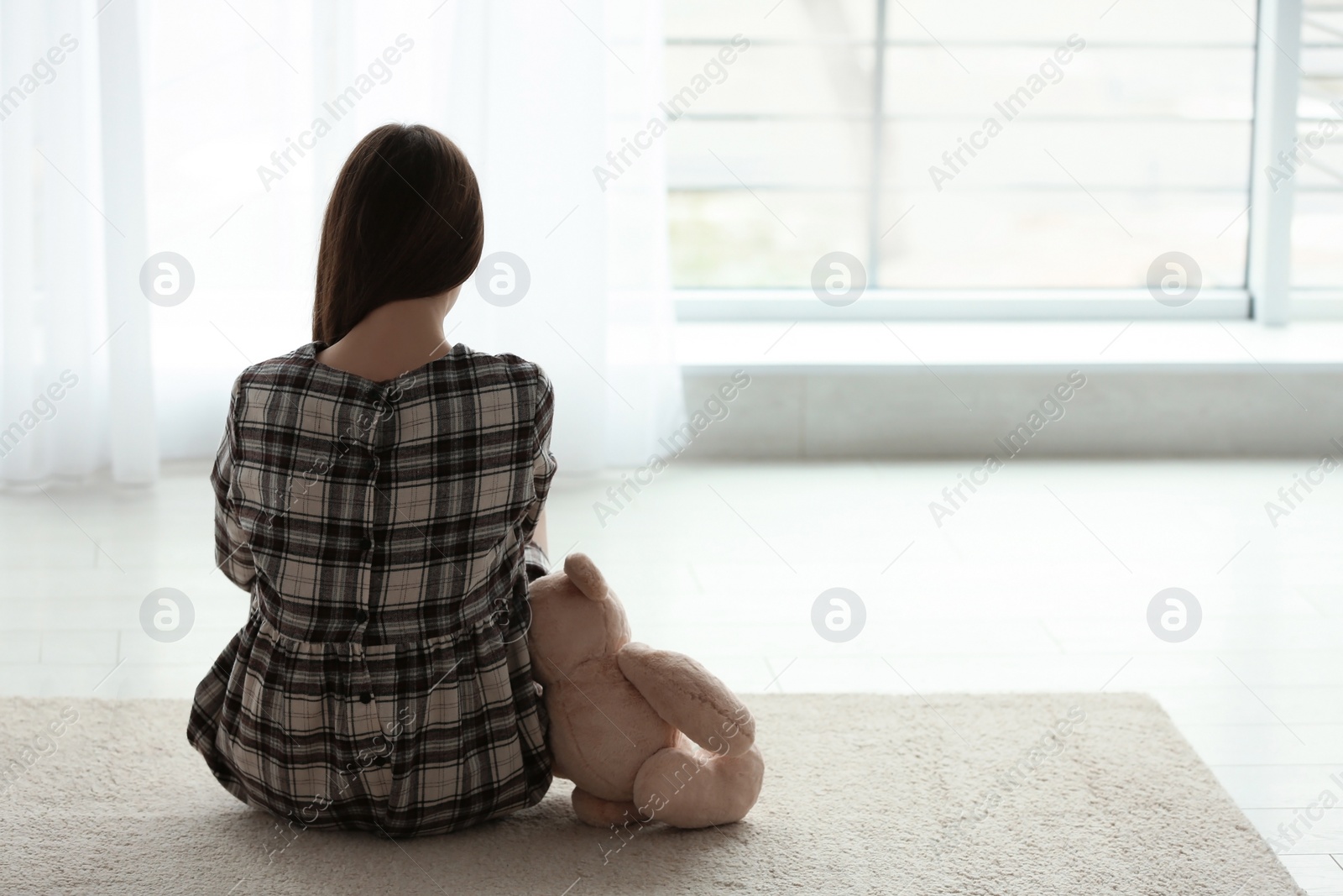 Photo of Upset girl with toy sitting near window indoors. Space for text