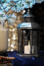 Photo of Arabic lantern, misbaha and vase with flowers on table against blurred lights