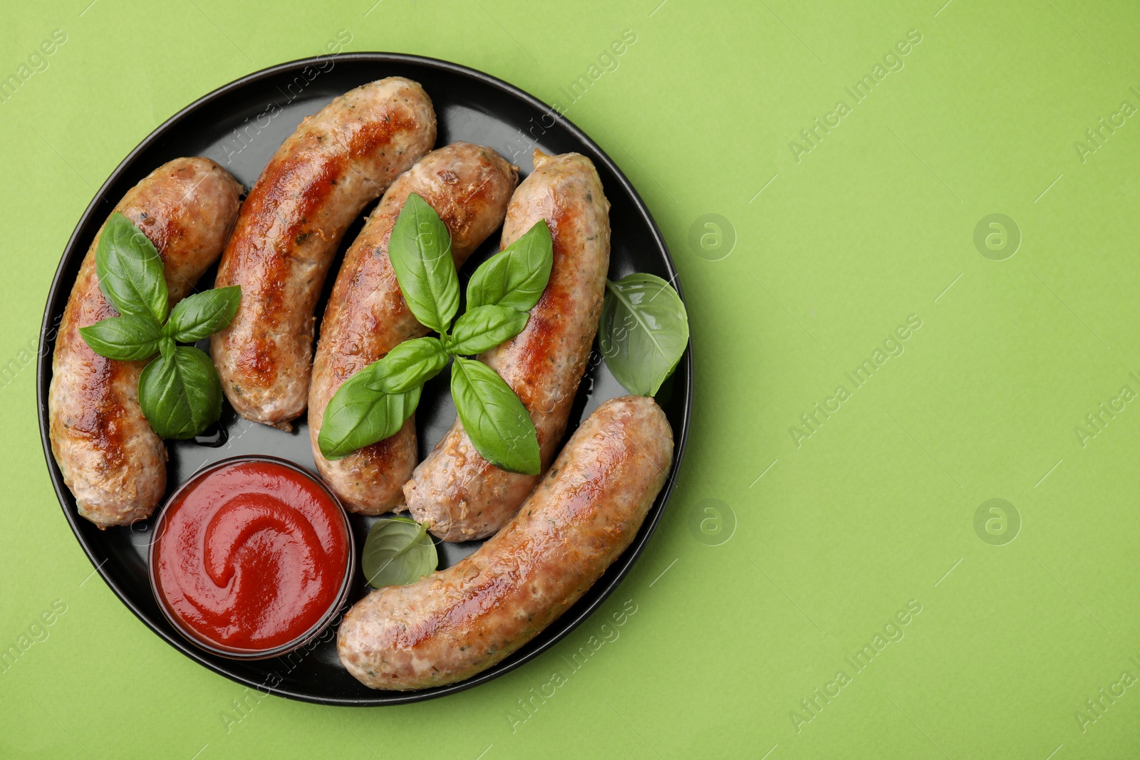 Photo of Plate with tasty homemade sausages, ketchup and basil leaves on green table, top view. Space for text