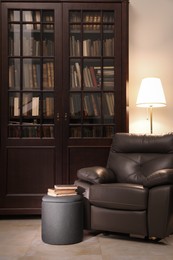 Photo of Comfortable armchair with pouf, books and lamp near wooden bookcase in library