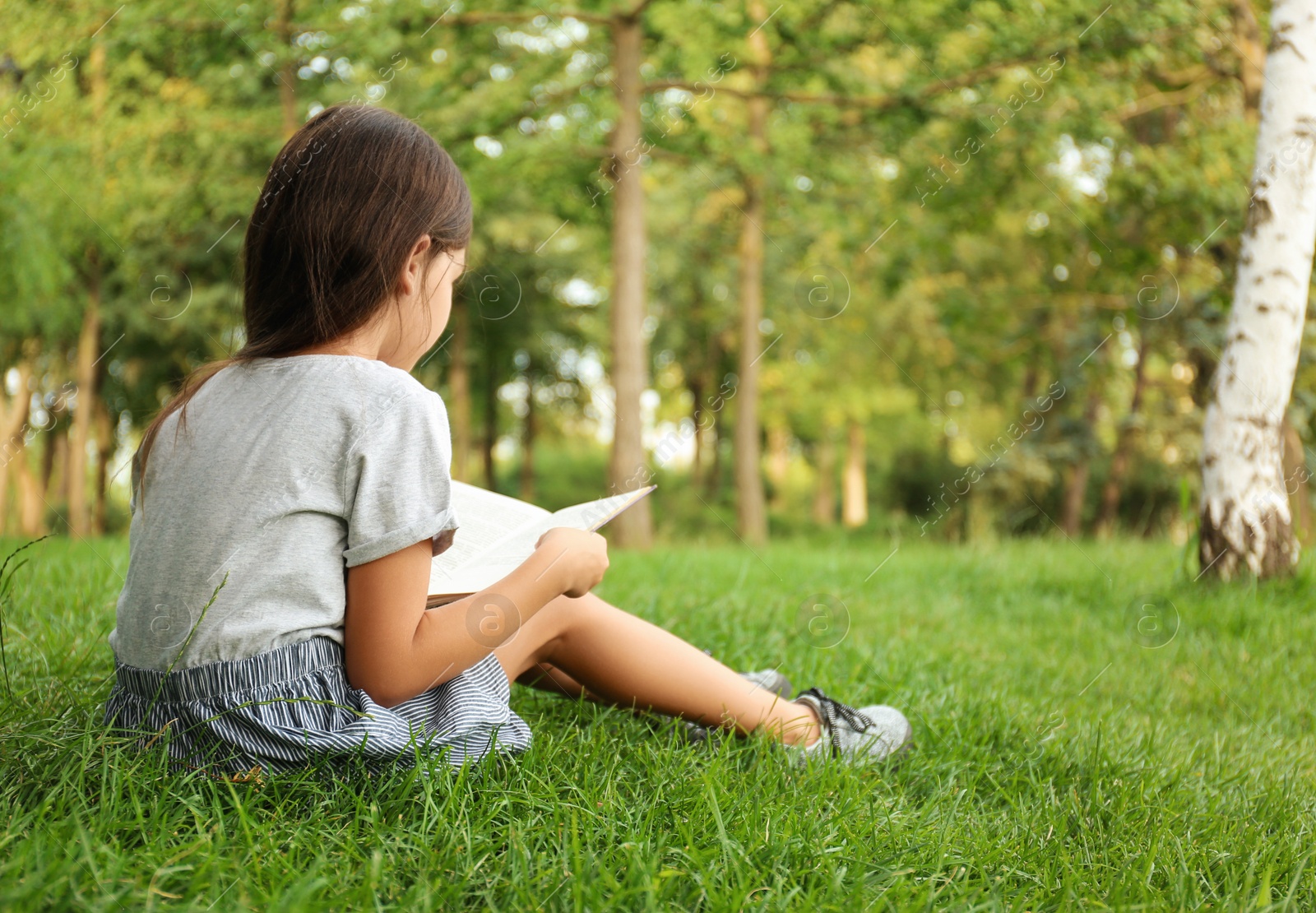 Photo of Cute little girl reading book on green grass in park