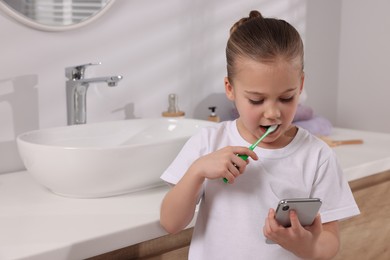 Photo of Little girl using smartphone while brushing teeth in bathroom, space for text. Internet addiction