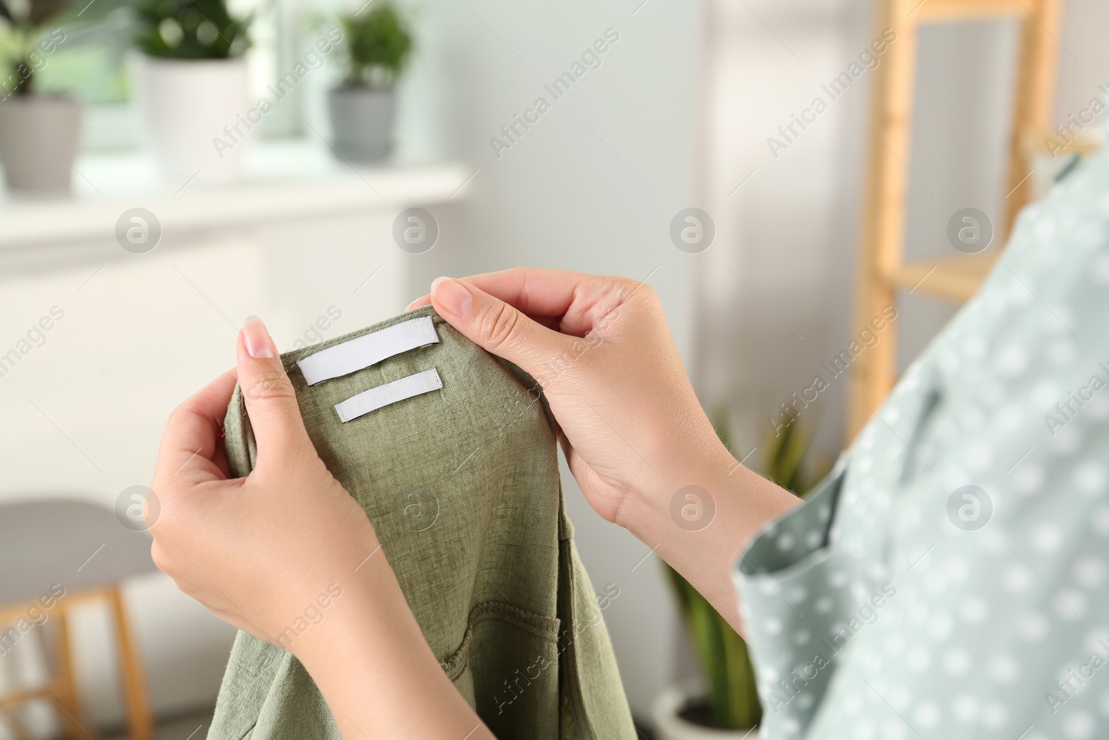 Photo of Woman holding green apparel with blank clothing label at home, closeup