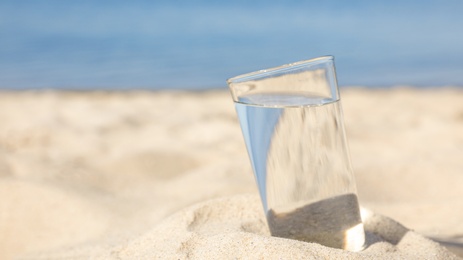 Photo of Sandy beach with glass of refreshing drink on hot summer day, space for text