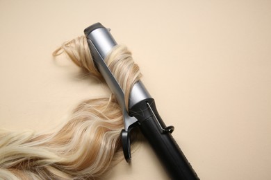 Curling iron with blonde hair lock on beige background. Space for text