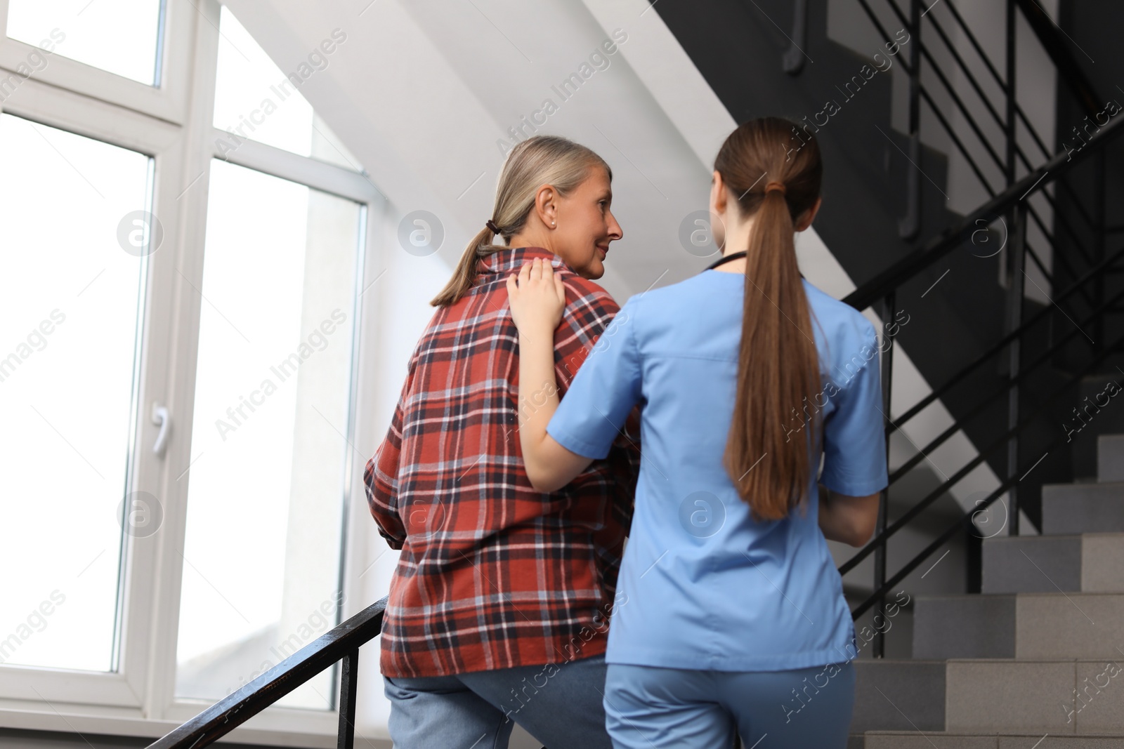 Photo of Young healthcare worker assisting senior woman on stairs indoors, low angle view