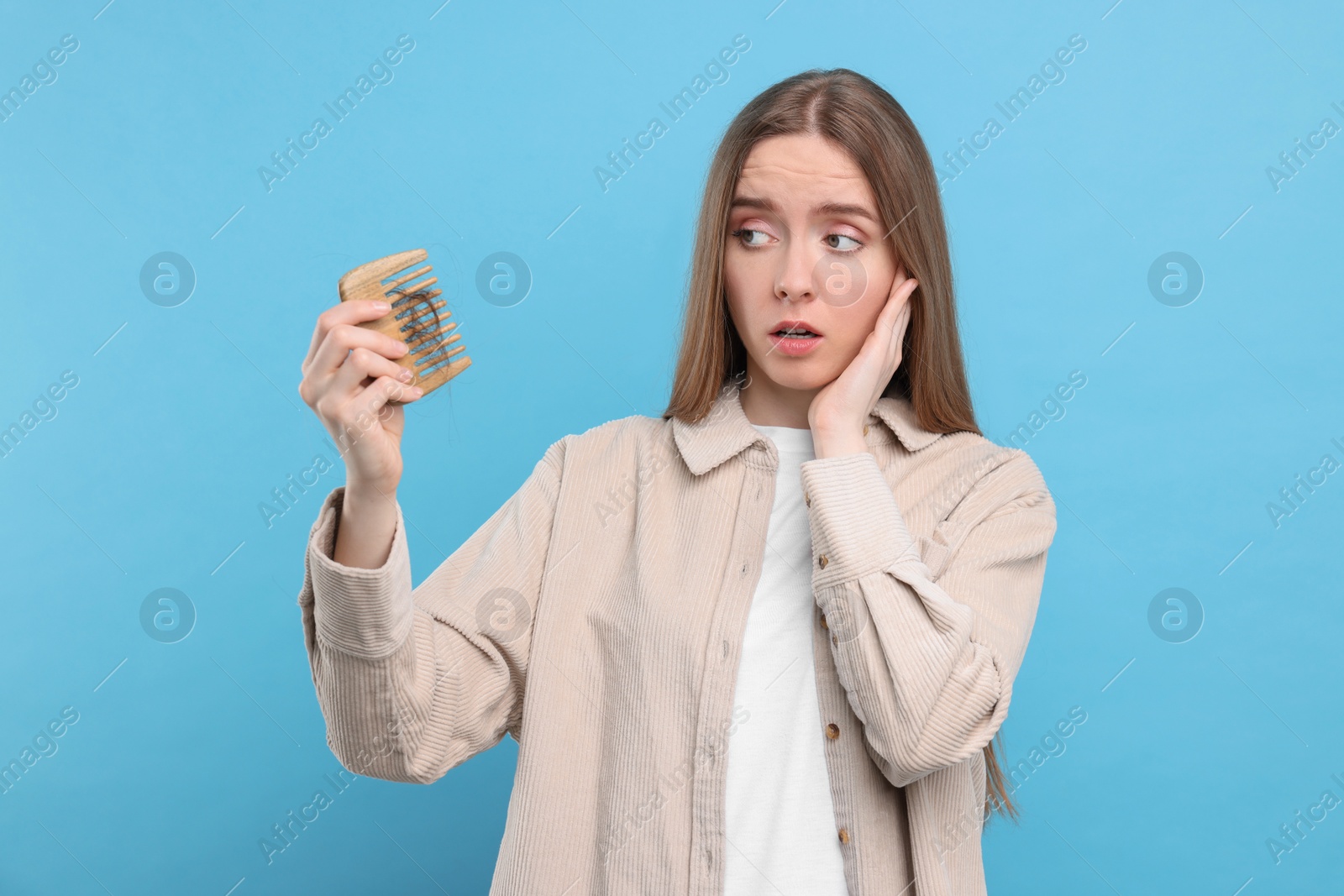 Photo of Emotional woman holding comb with lost hair on light blue background. Alopecia problem