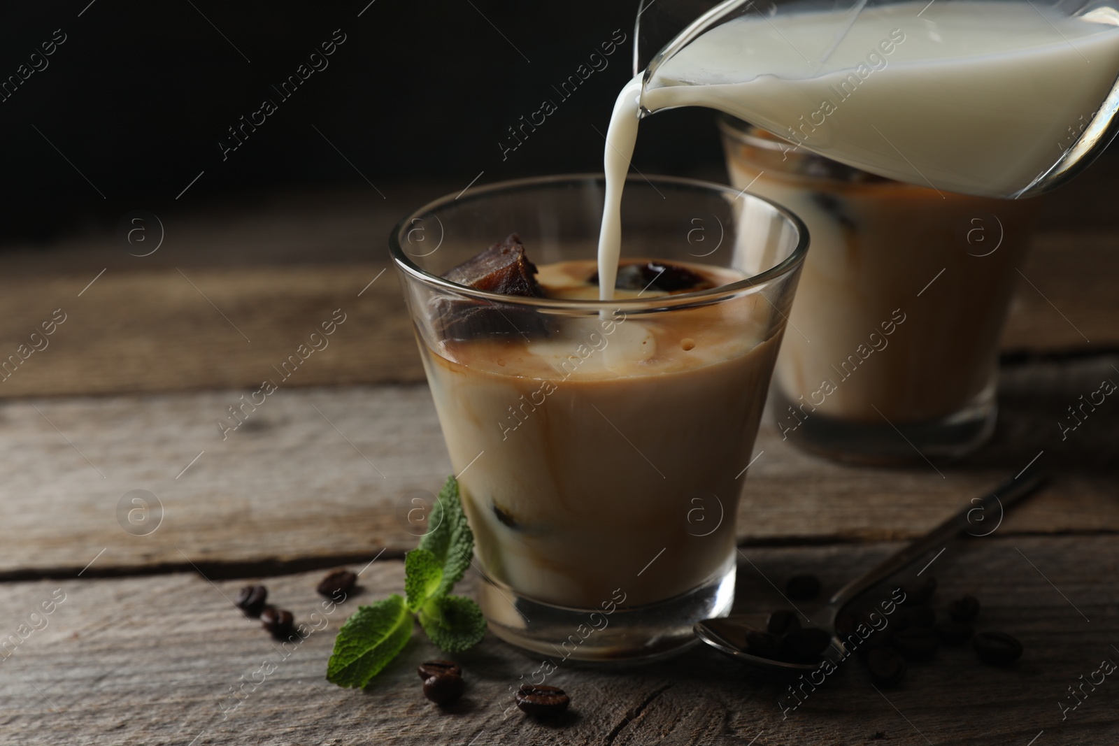 Photo of Pouring milk into glass of delicious iced coffee, mint and beans on wooden table, closeup
