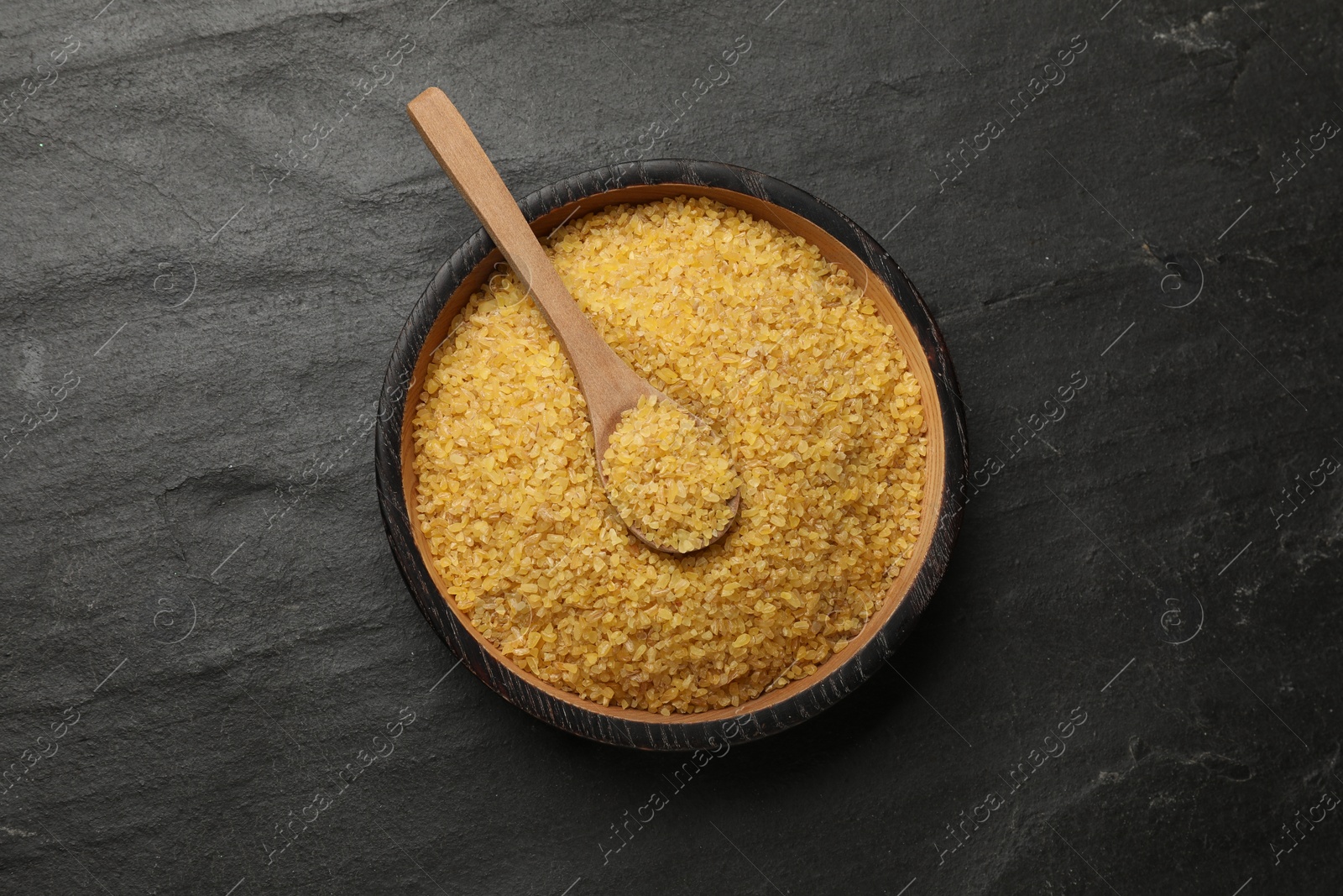 Photo of Bowl and spoon with raw bulgur on dark gray table, top view
