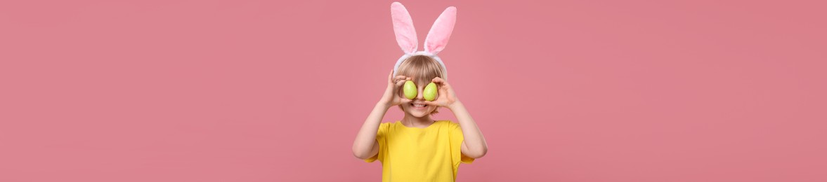 Image of Happy child with bunny ears holding Easter eggs near eyes on pink background. Banner design