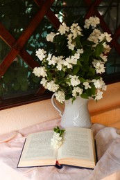 Photo of Bouquet of beautiful jasmine flowers in vase and open book on fabric indoors