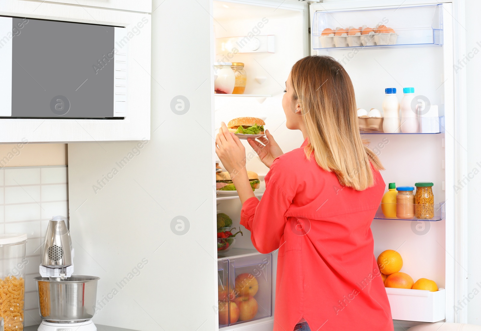 Photo of Young woman taking sandwich from refrigerator in kitchen. Failed diet