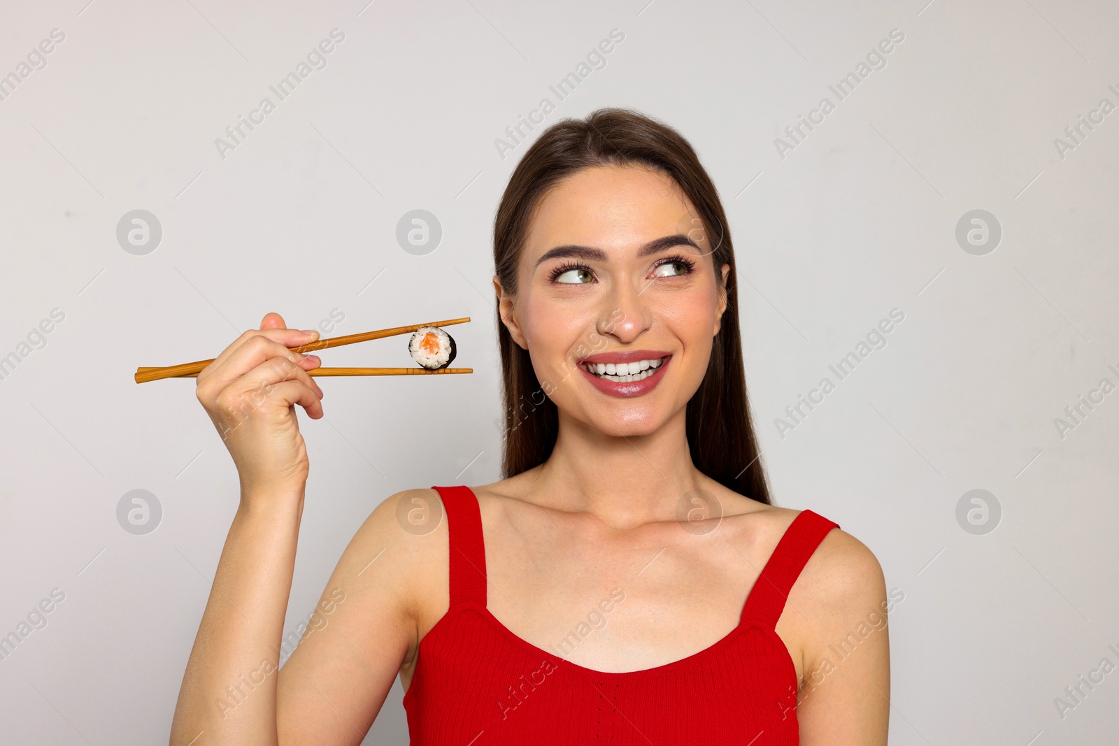 Photo of Happy beautiful young woman holding sushi roll with chopsticks on light background