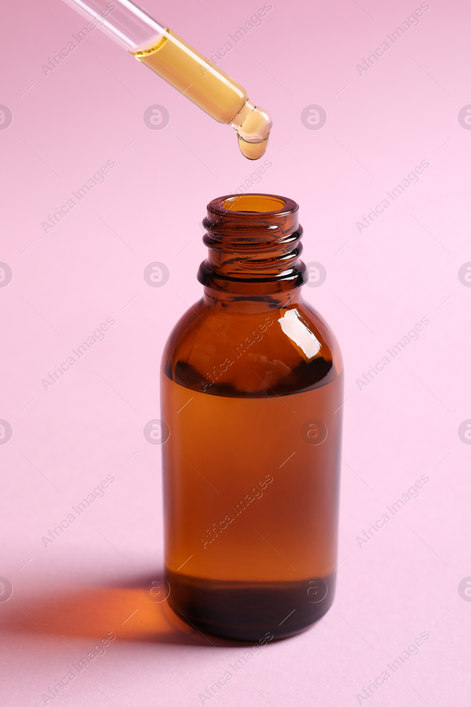 Photo of Dripping cosmetic oil from pipette into bottle on pink background