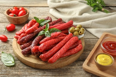 Photo of Different thin dry smoked sausages, basil, sauces and olives on wooden table