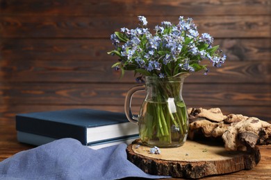Photo of Bouquet of beautiful forget-me-not flowers in glass jug, books and piece of decorative wood on wooden table, space for text