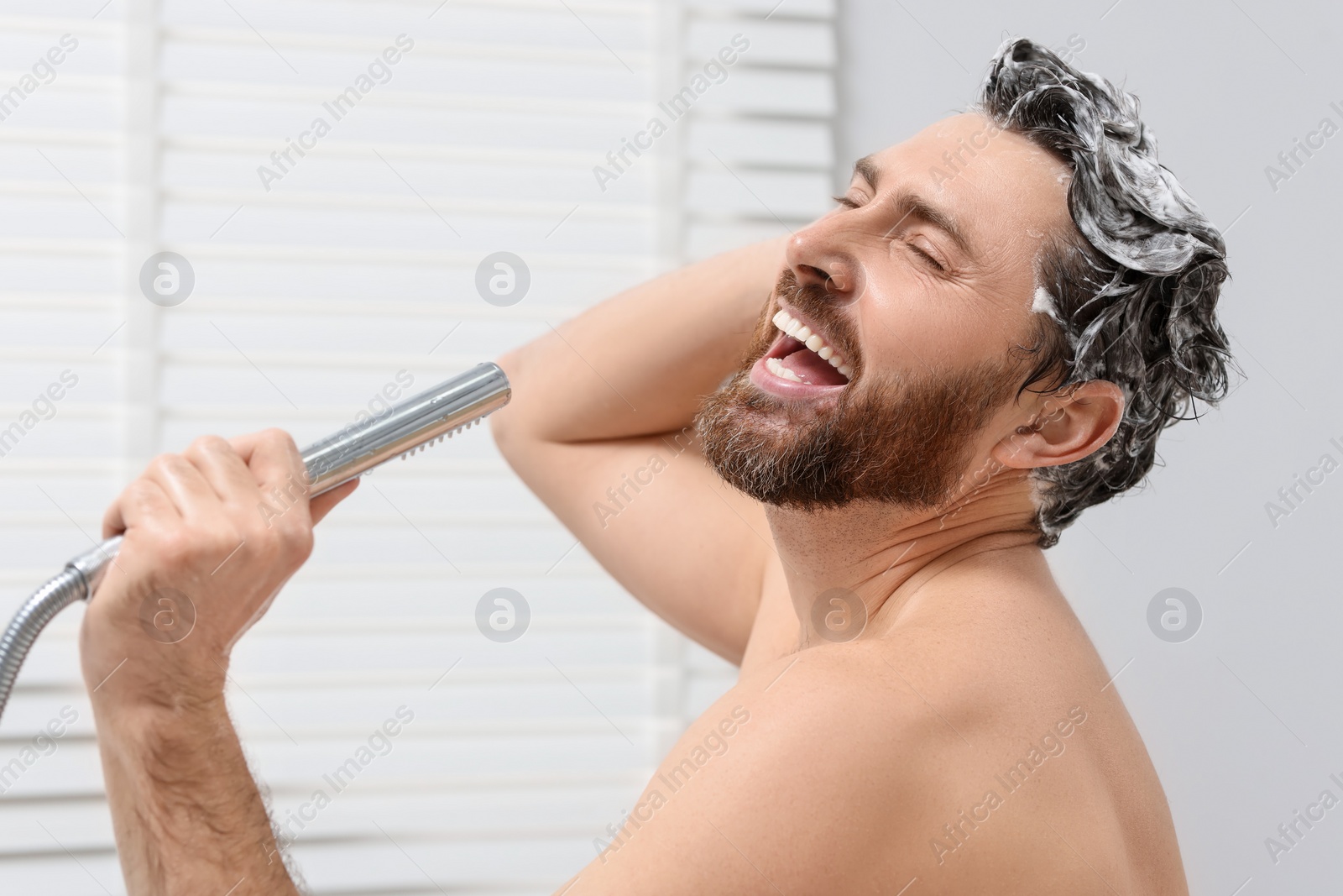 Photo of Happy man with showerhead singing and washing his hair with shampoo indoors