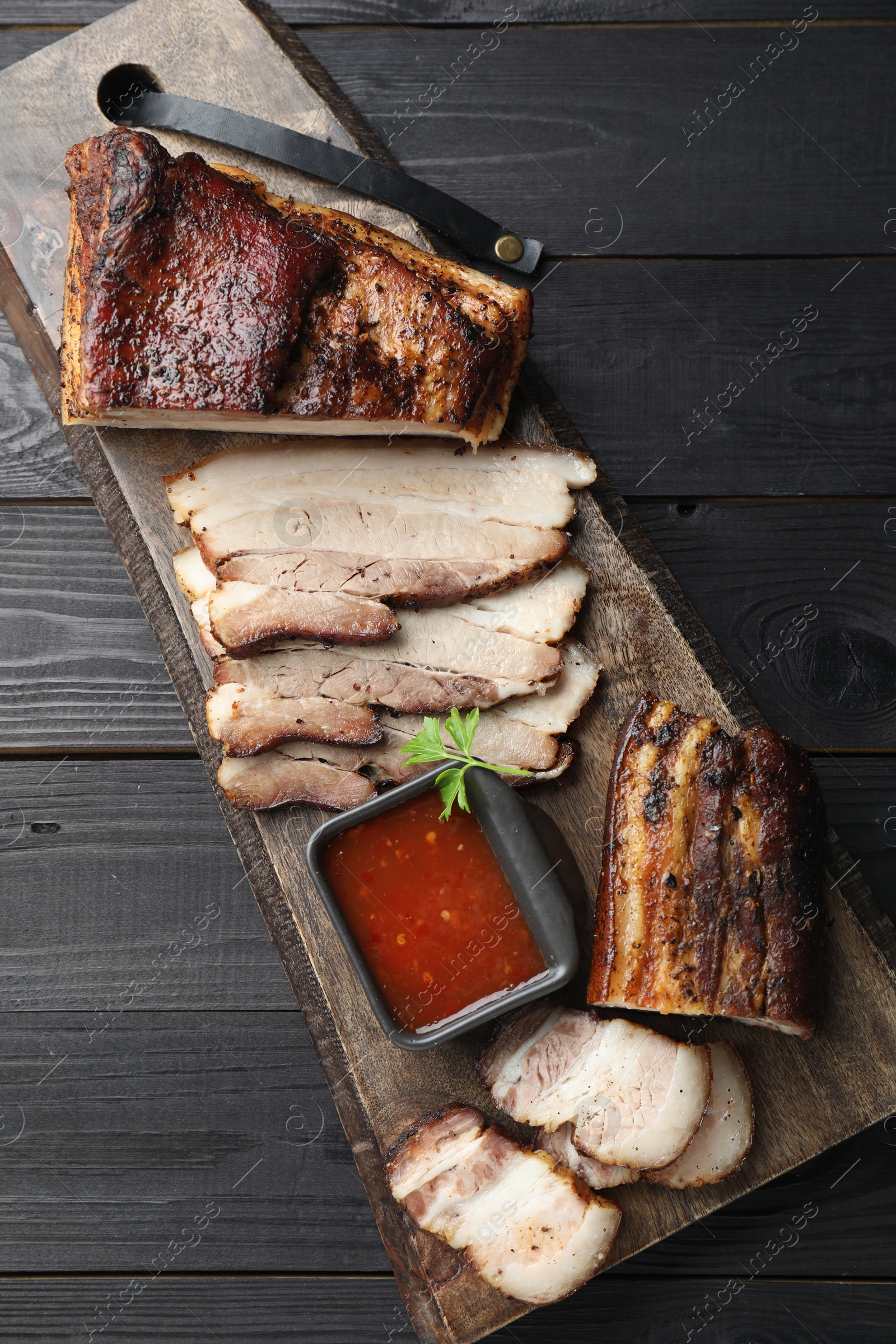 Photo of Pieces of baked pork belly served with sauce and parsley on black wooden table, top view