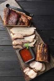 Photo of Pieces of baked pork belly served with sauce and parsley on black wooden table, top view