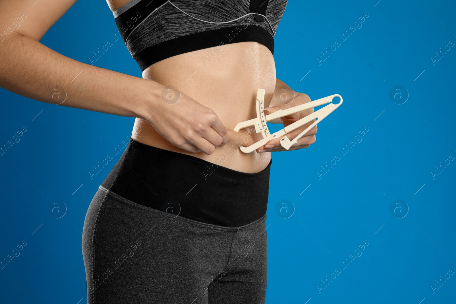 Photo of Young woman measuring body fat with caliper on blue background, closeup