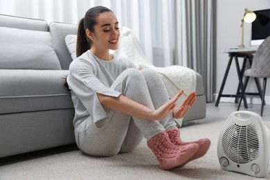 Photo of Young woman warming hands near electric heater at home