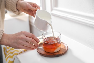 Woman pouring milk into cup of tea at white table, closeup