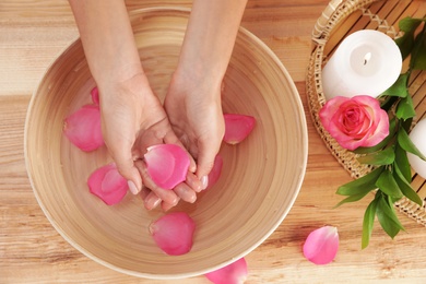 Photo of Woman soaking her hands in bowl with water and petals on wooden table, top view. Spa treatment