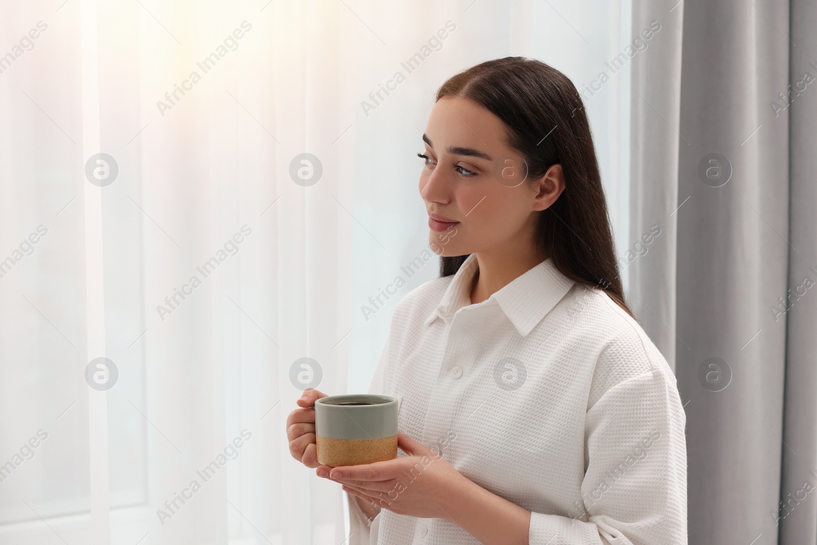 Photo of Beautiful woman with cup of drink near window at home, space for text. Lazy morning