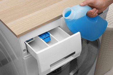 Photo of Man pouring fabric softener from bottle into washing machine indoors, closeup