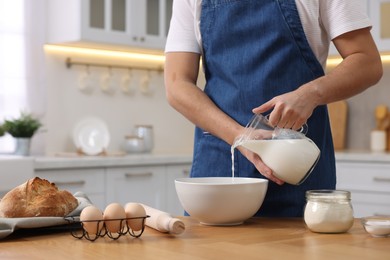 Photo of Making bread. Man pouring milk into bowl at wooden table in kitchen, closeup