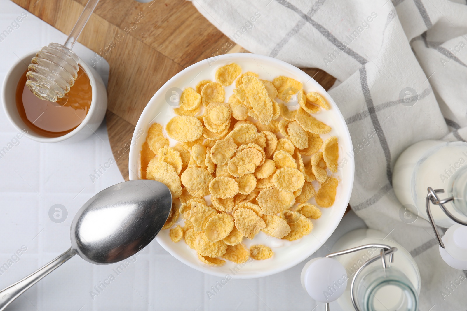 Photo of Breakfast cereal. Tasty corn flakes, milk, honey and spoon on white tiled table, top view