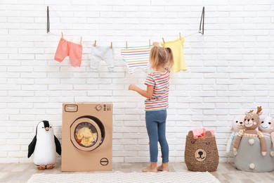 Photo of Little girl hanging clean laundry near toy cardboard washing machine indoors