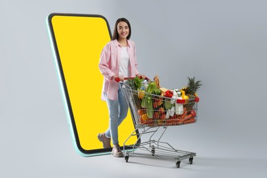 Image of Grocery shopping via internet. Happy woman with shopping cart full of products walking out of huge smartphone on grey background