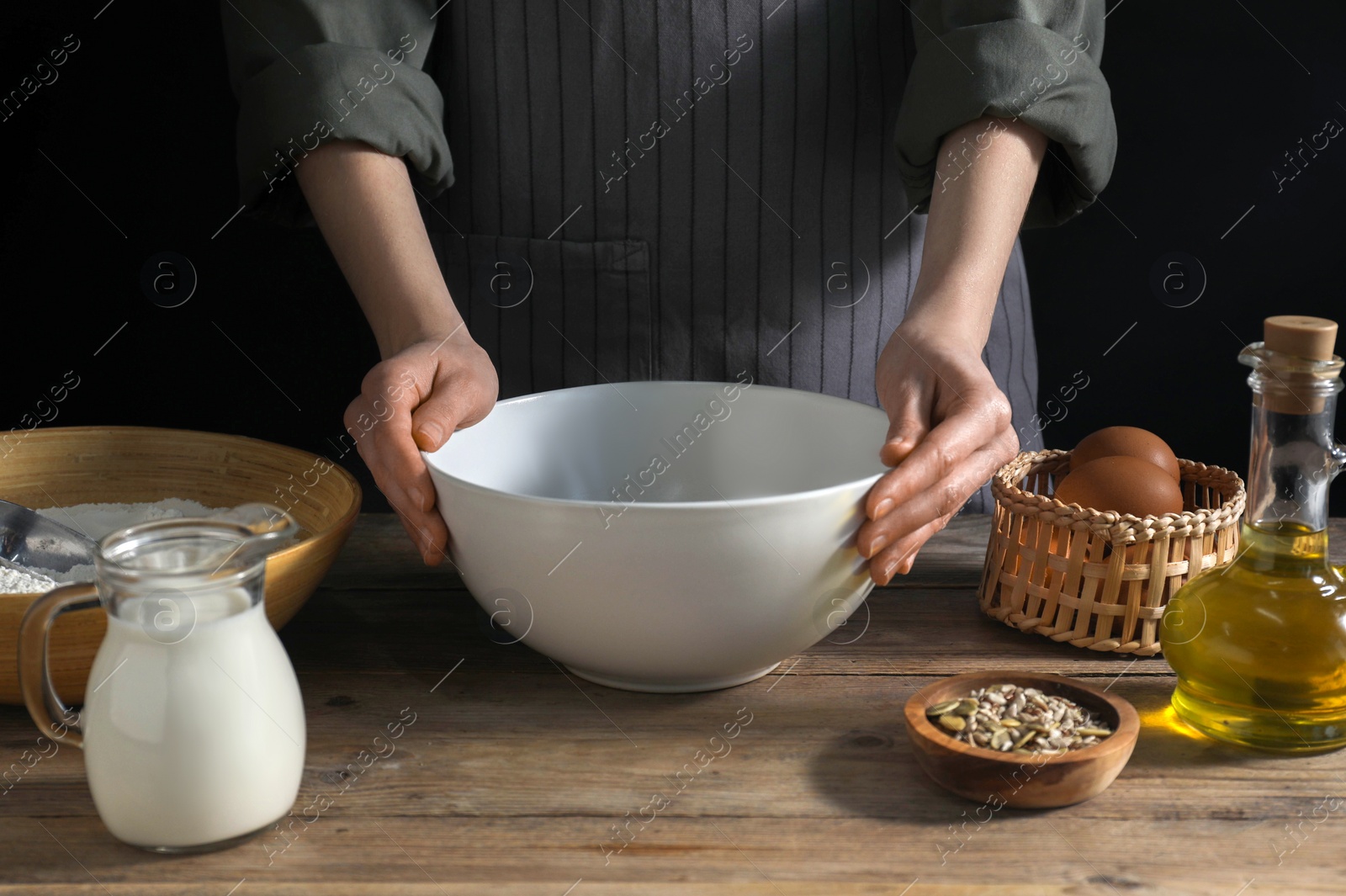 Photo of Making bread. Woman with ingredients for dough at wooden table on dark background, closeup