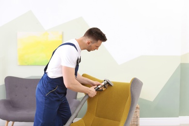 Photo of Male worker removing dirt from armchair with professional vacuum cleaner indoors