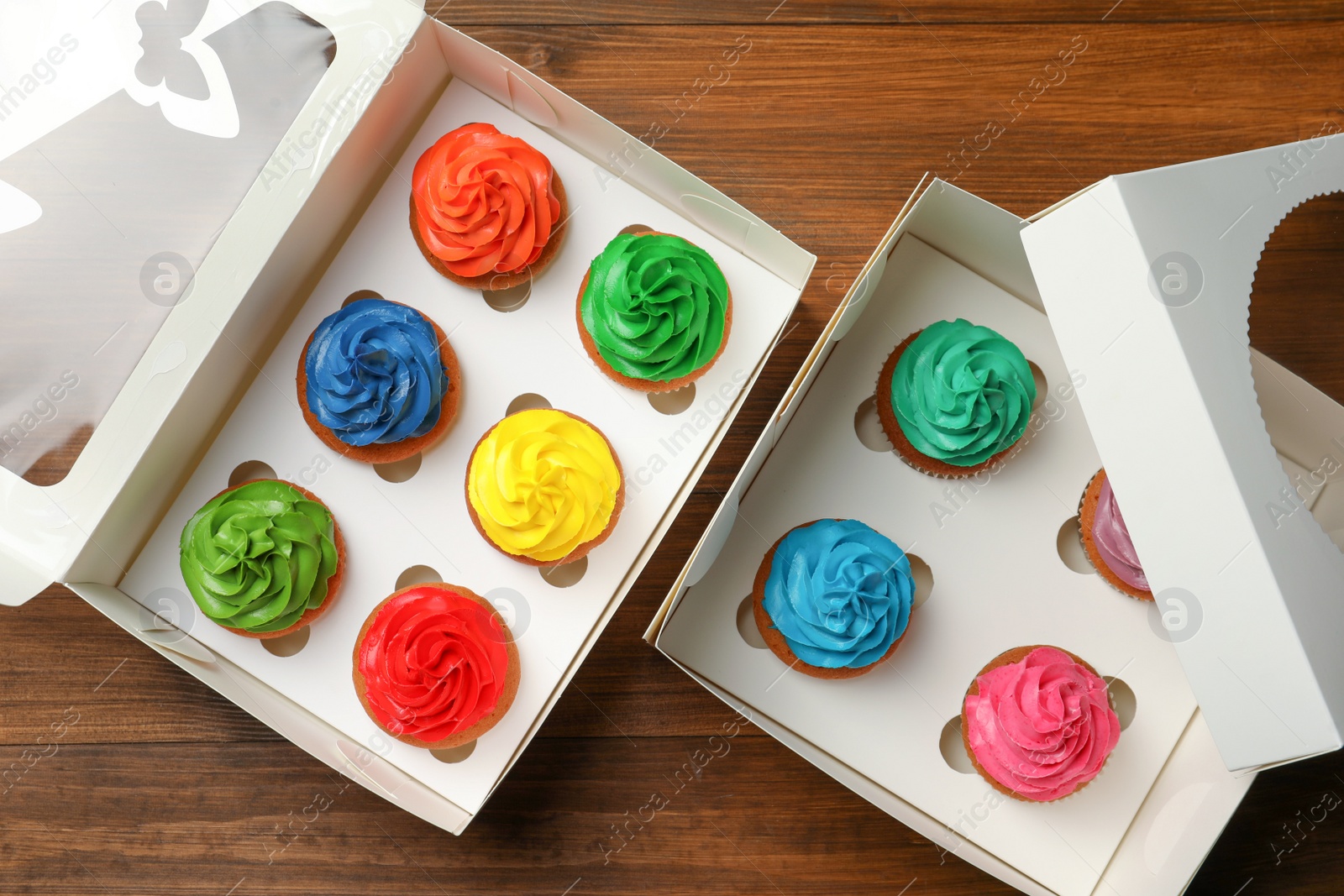 Photo of Boxes with delicious colorful cupcakes on wooden table, flat lay