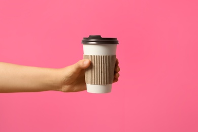 Woman holding takeaway paper coffee cup with cardboard sleeve on pink background, closeup