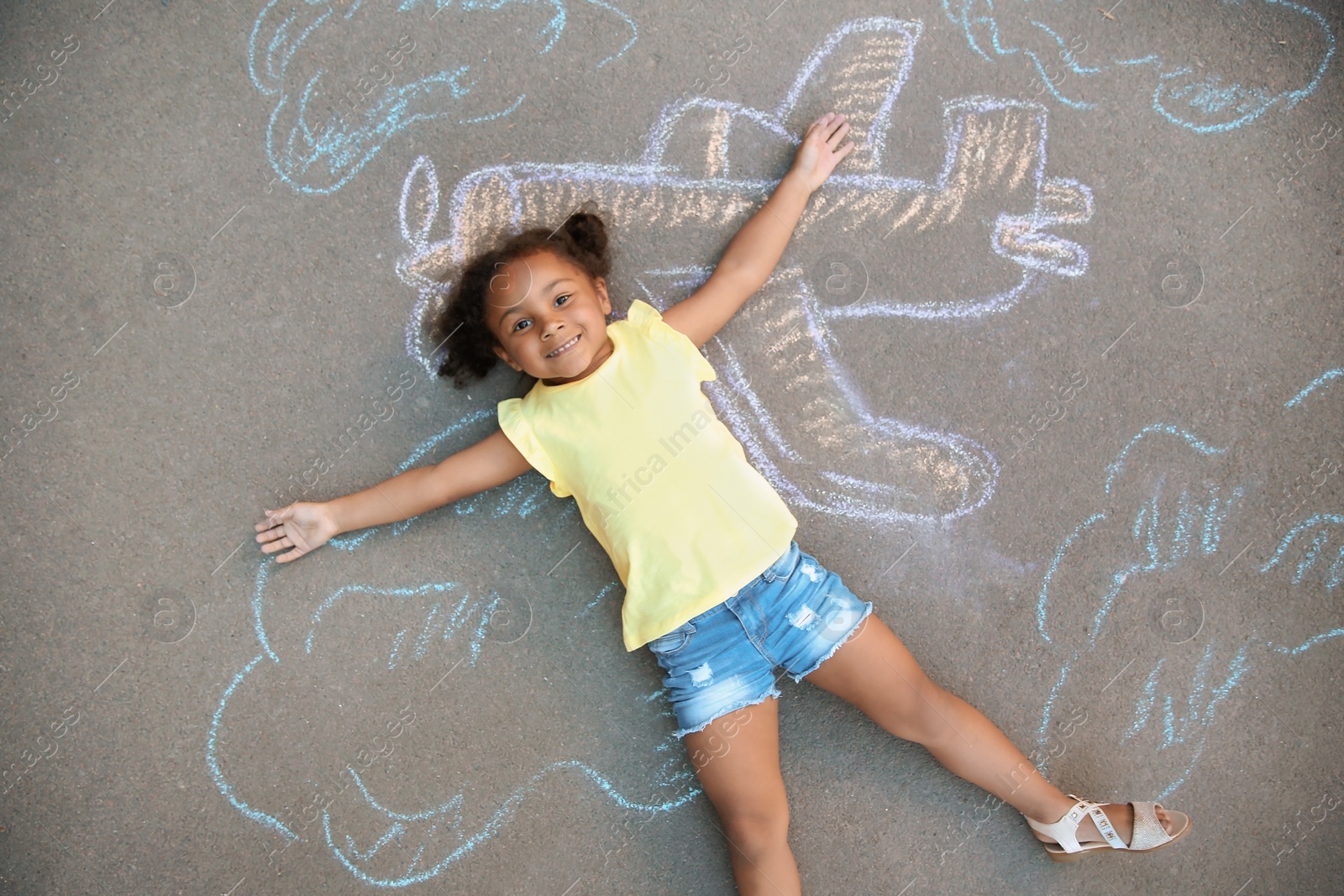 Photo of Little African-American child lying near chalk drawing of airplane on asphalt, top view