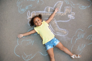 Little African-American child lying near chalk drawing of airplane on asphalt, top view