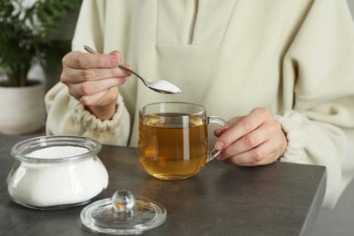 Woman adding sugar into aromatic tea at grey table, closeup