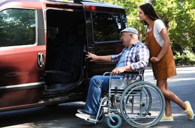 Photo of Young woman helping senior man in wheelchair to get into van outdoors