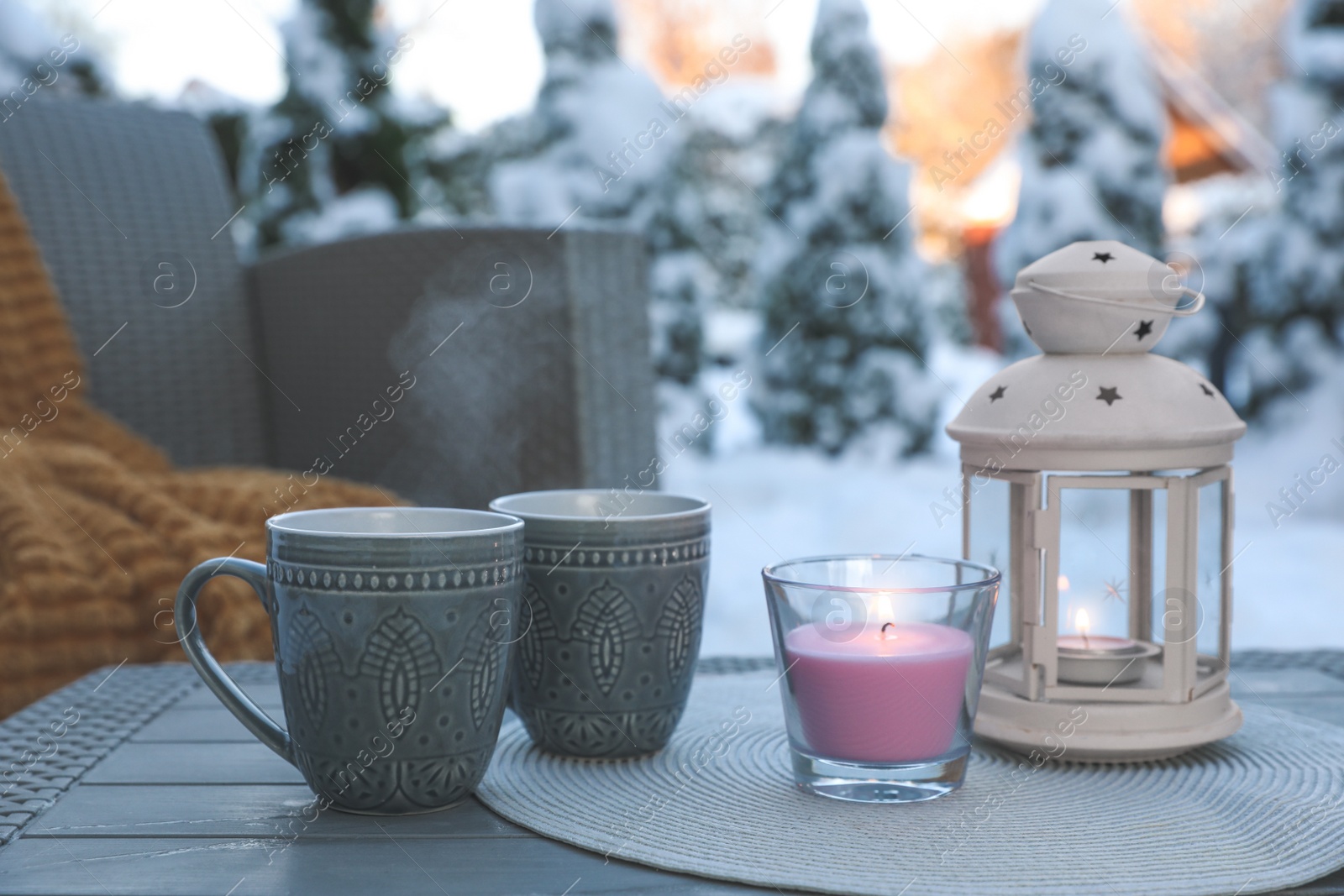 Photo of Burning candle, lantern and cups with hot drink on coffee table outdoors. Cosy winter