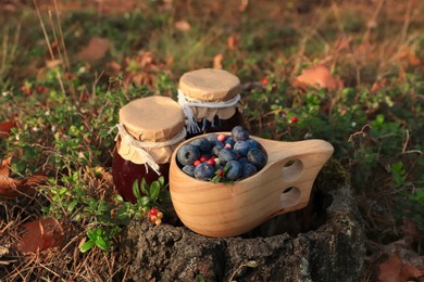 Wooden mug full of fresh ripe blueberries, lingonberries and jars with jam on stump