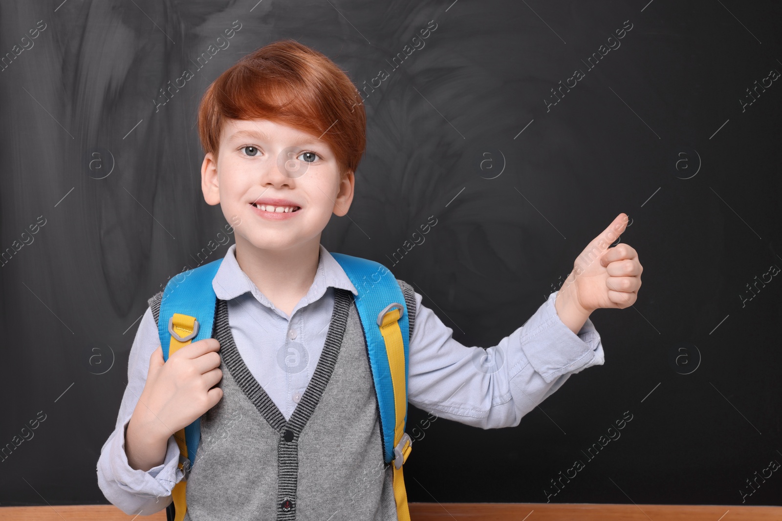 Photo of Smiling schoolboy showing thumb up near blackboard