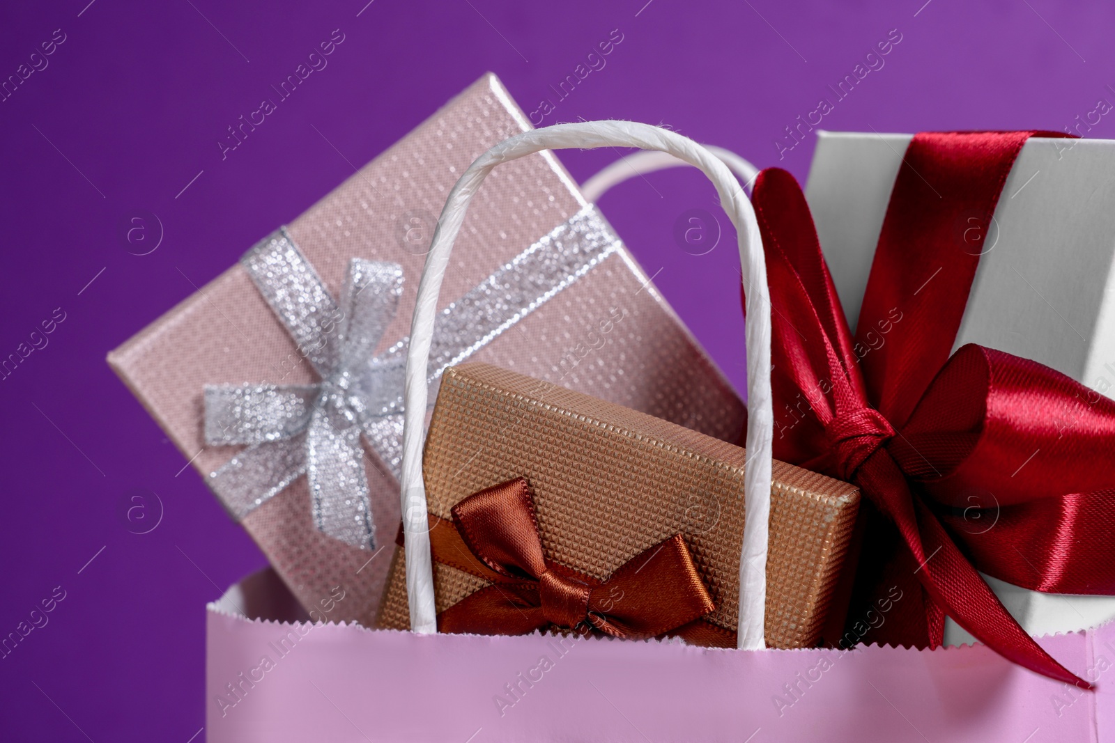 Photo of Pink paper shopping bag full of gift boxes on purple background, closeup