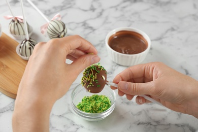 Young woman decorating tasty cake pop with green sprinkles at white marble table, closeup